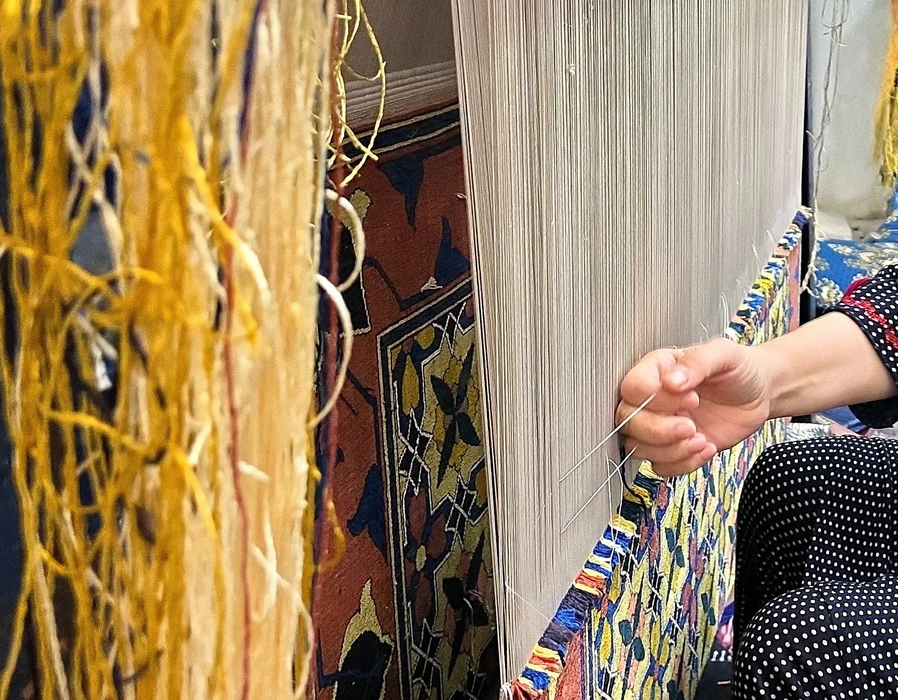 Mains d'une tisserande passant un fil sur un métier à tisser, devant un tapis aux motifs colorés